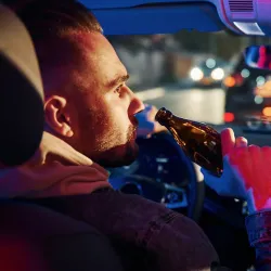 A profile view of a person drinking from a beer bottle while sitting in the driver's seat of a car at night. The image is lit with blue and red ambient lighting, and street lights can be seen blurred in the background through the windshield. This image depicts dangerous and illegal behavior of drinking while driving.